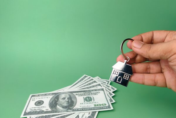 A hand holding a house-shaped keychain above hundred-dollar bills.
