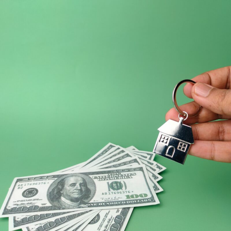 A hand holding a house-shaped keychain above hundred-dollar bills.