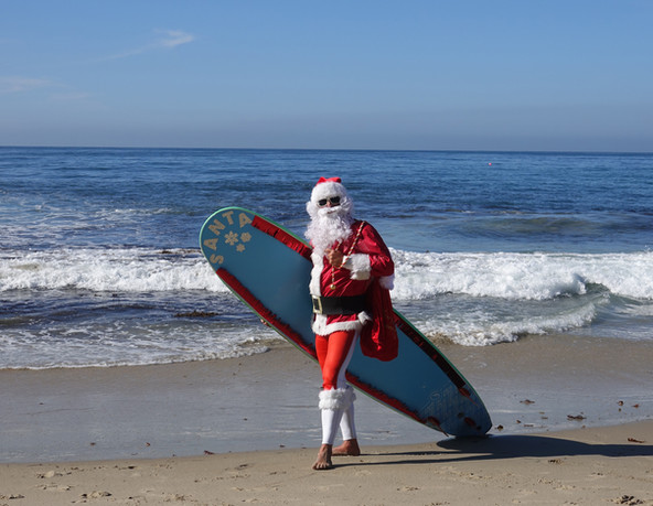 A Person Dressed as Santa in Festive Red Suit with White Trim Holding a Surfboard by The Ocean